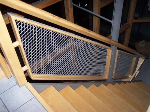 Expanded metal infill panels with galvanized surface and diamond holes are installed on the wooden stair railing indoors.